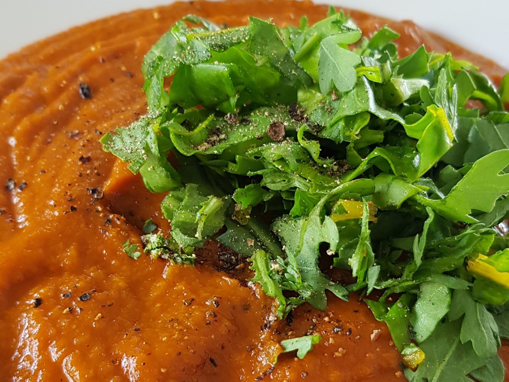 Deep red-orange soup close up with chopped green leaves on top, and black pepper